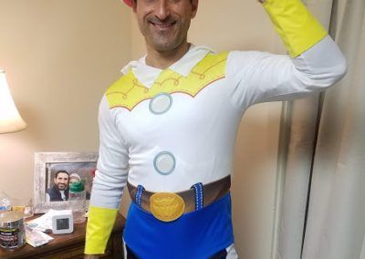 A person wears a Woody from Toy Story costume, smiling and holding one arm up in a pose in an indoor room.