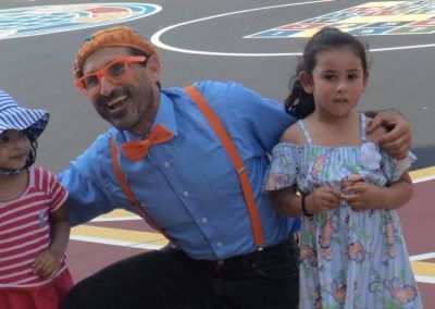 A smiling person in a blue shirt, orange suspenders, and bow tie poses for a photo between two children outdoors.