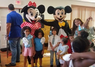 People pose with Mickey and Minnie Mouse mascots at an indoor party, with some guests wearing colorful party hats.