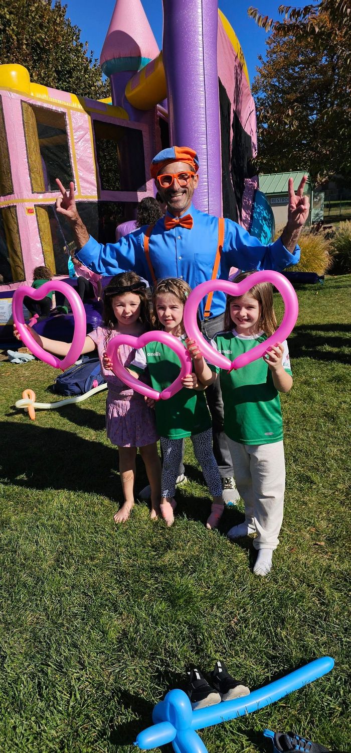 An entertainer in a blue outfit and children holding balloon hearts pose outdoors in front of an inflatable castle.
