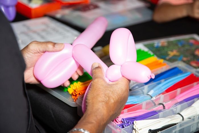 Hands shape pink modeling balloons, with a container of assorted colored balloons in the background.