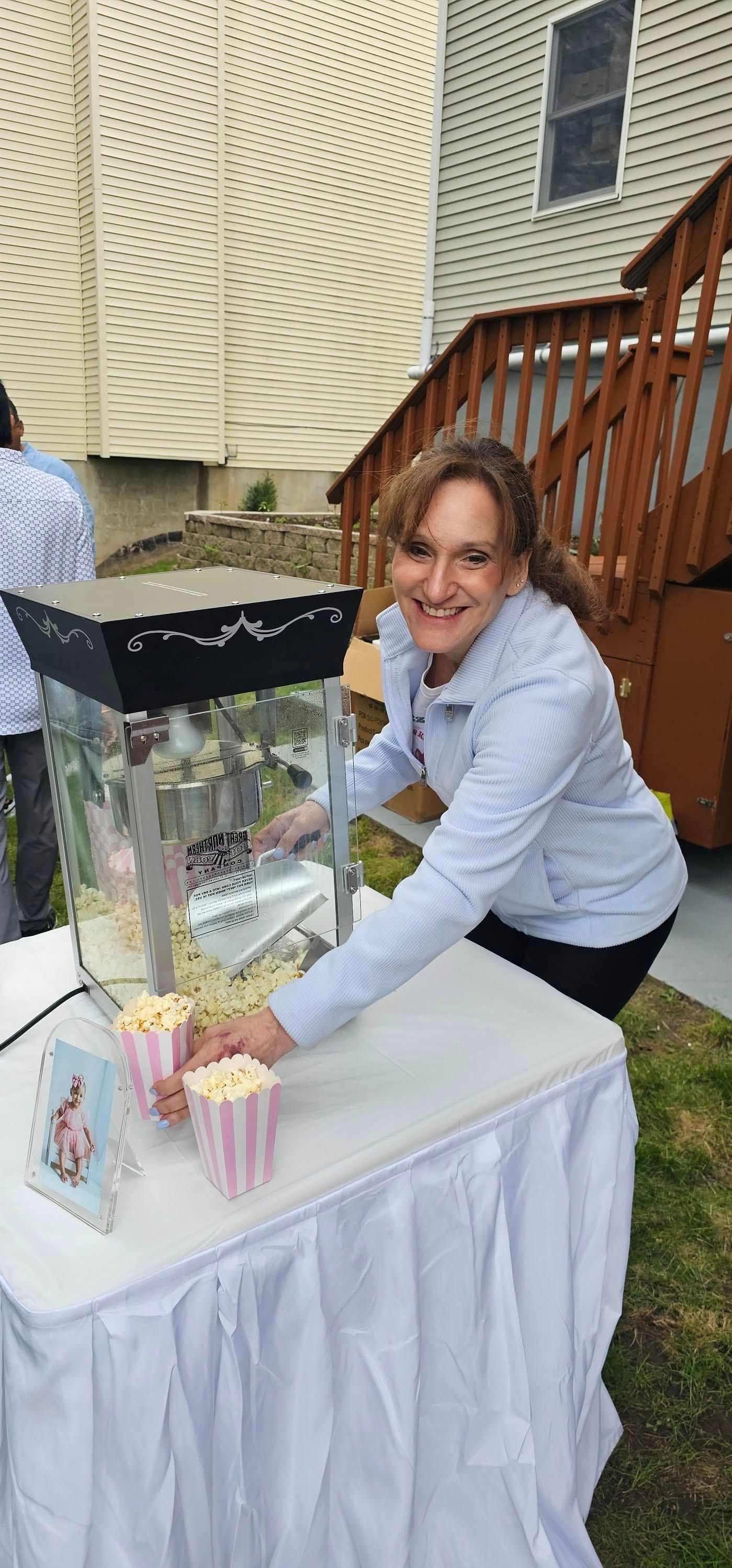 A smiling person in a light blue jacket stands at an outdoor table, serving popcorn from a machine into striped bags.