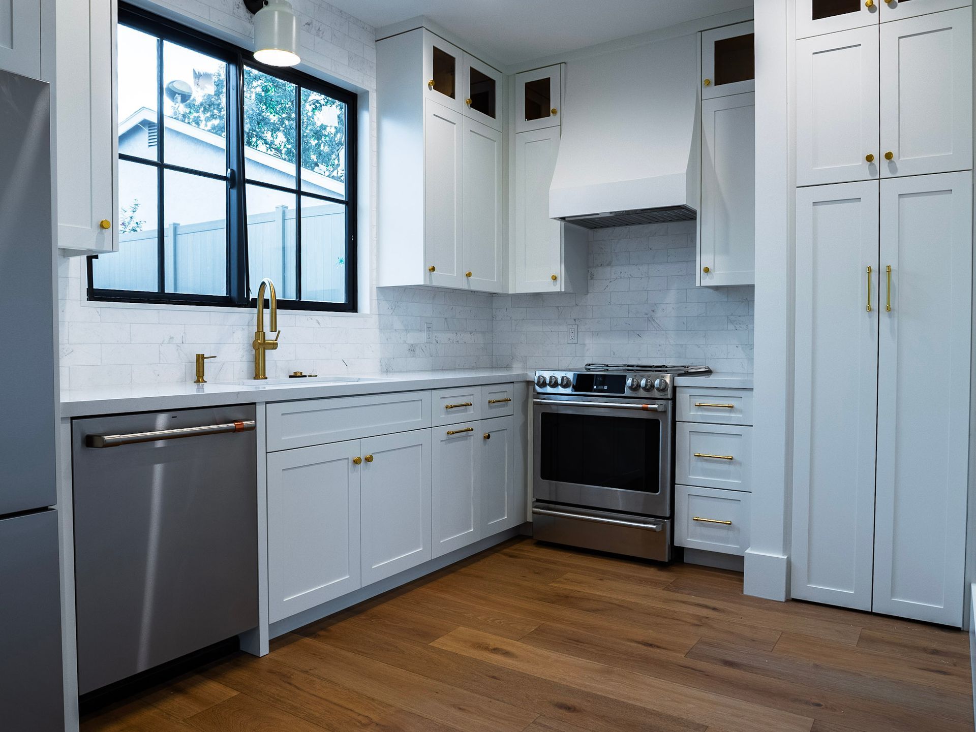 Modern white kitchen with stainless steel appliances, gold hardware, and wood floors.