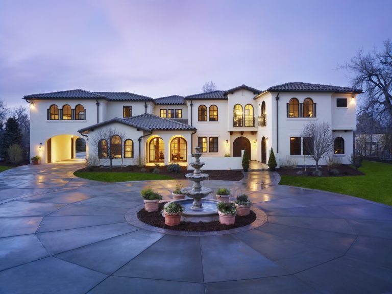 Large white Mediterranean-style mansion with circular driveway, fountain, and potted plants. Dusk lighting.