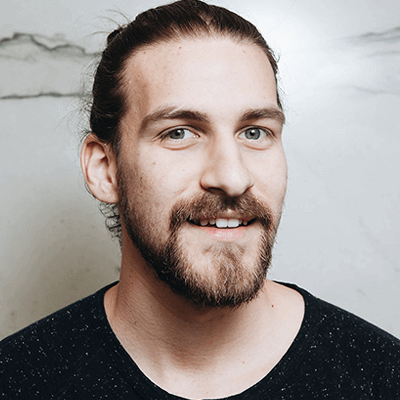 Homme avec une barbe souriant, cheveux tirés en arrière, chemise sombre, fond neutre.