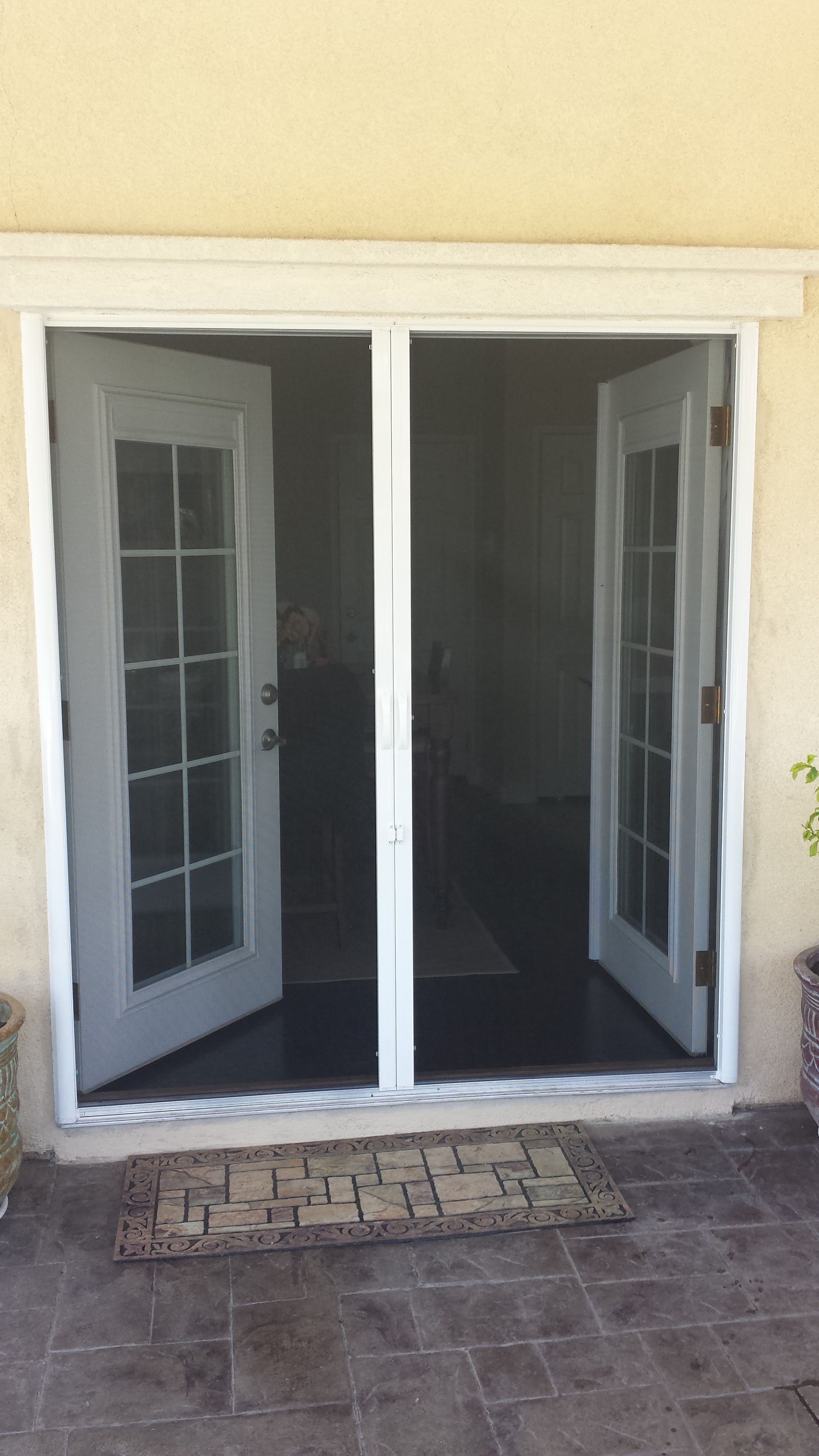 White double doors opened outward from a tan stucco building with a black mesh screen door installed in the entryway.
