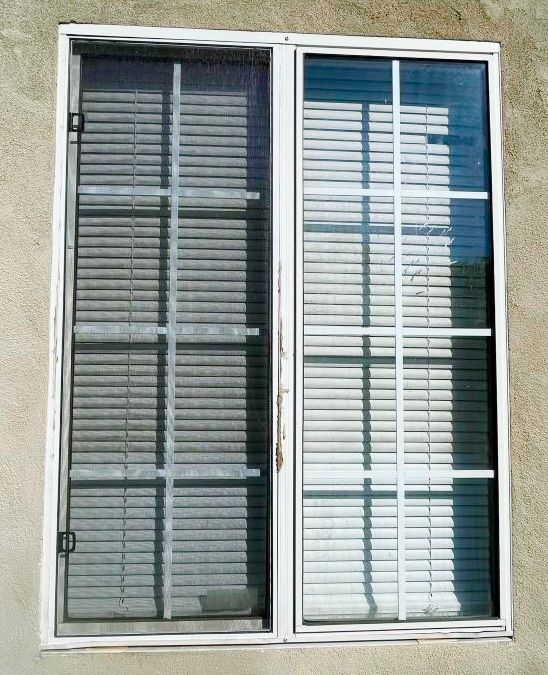 A rectangular window with a white frame and grid, featuring closed horizontal blinds set against a textured beige wall.