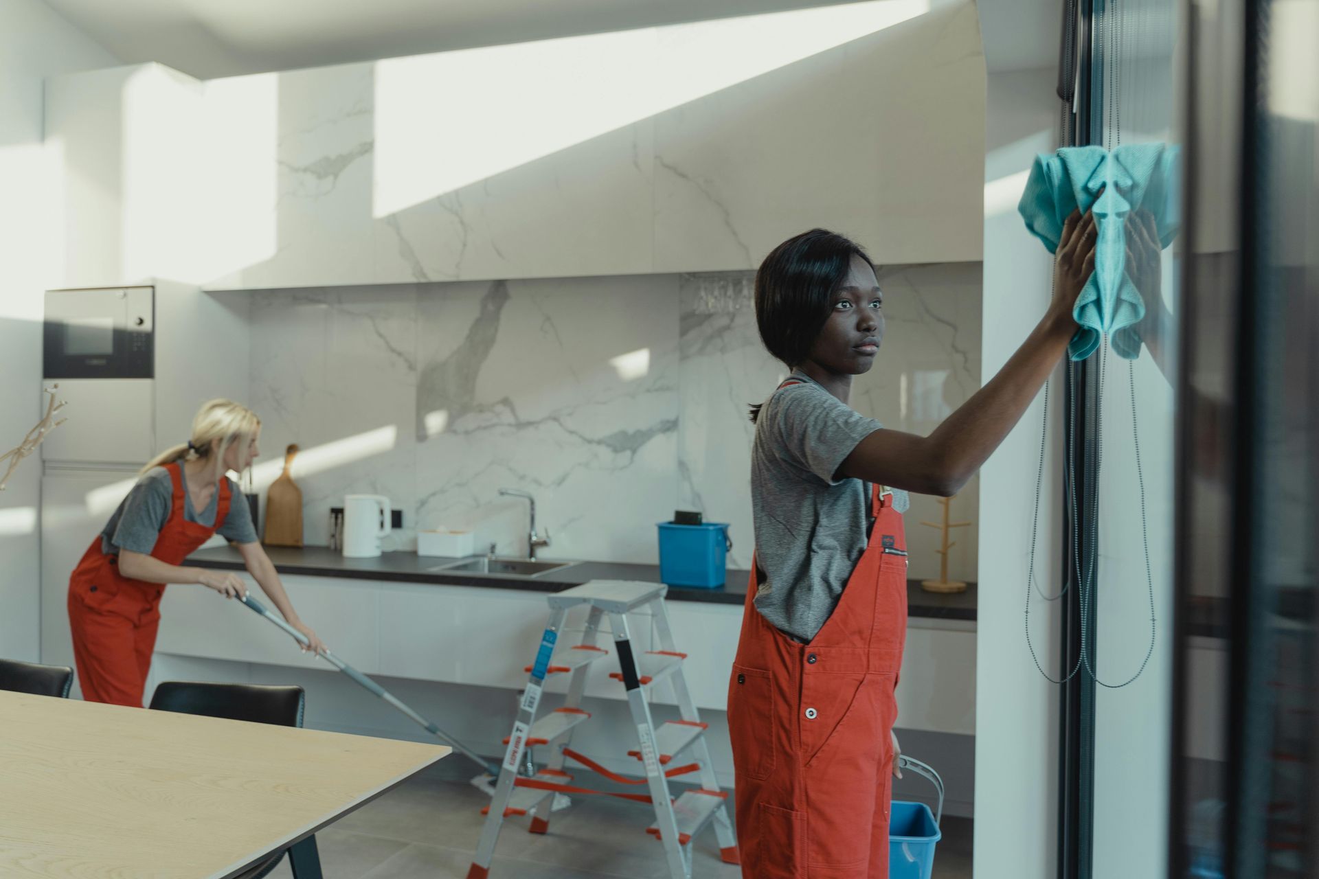 Two workers clean a bright modern kitchen, one wiping a glass wall and one mopping the floor.