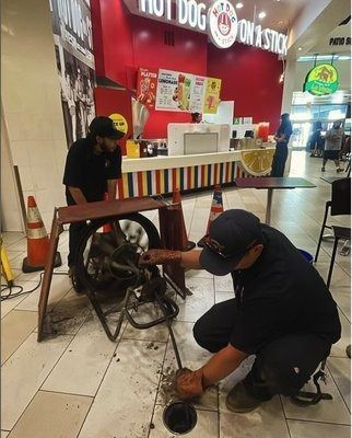 Two workers fix a drain in a mall food court. One kneels while the other stands. A 