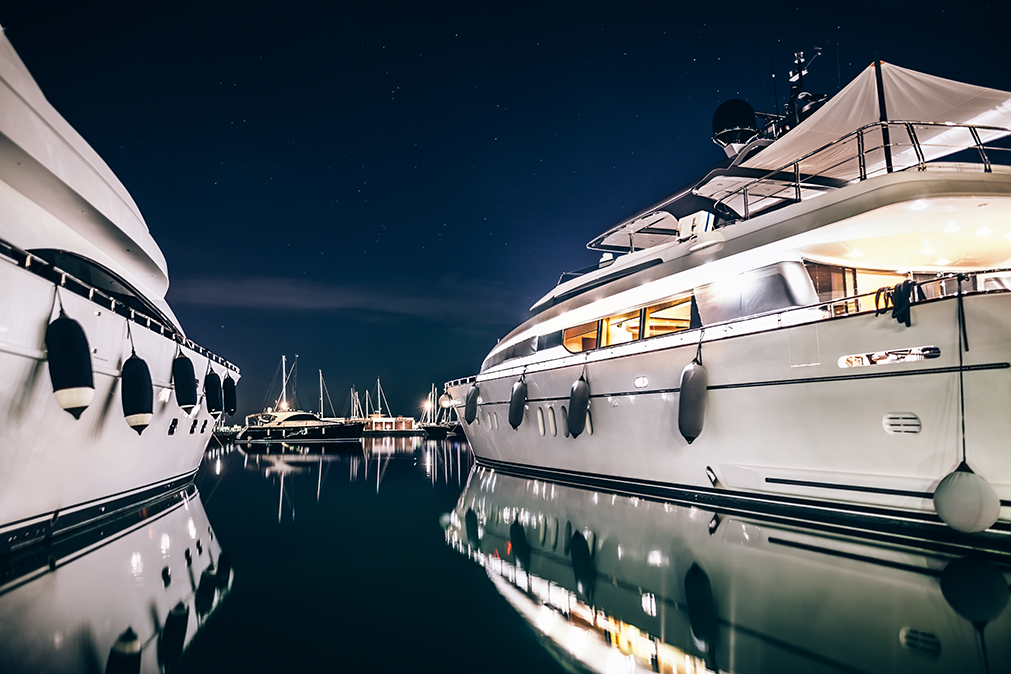 Two luxury yachts docked at night, reflecting on calm harbor water under city lights