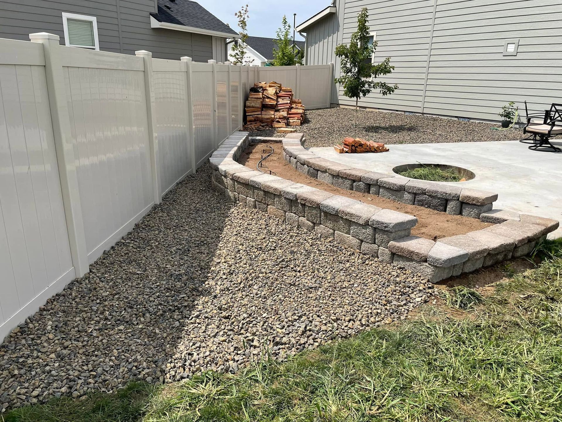 Backyard landscaping with curved stone retaining wall, gravel path, and stacked rocks beside a house and fence