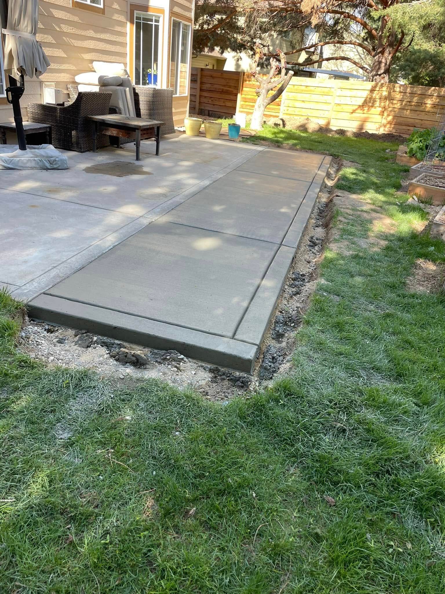 Concrete patio walkway beside a house, bordered by grass and outdoor furniture in a backyard