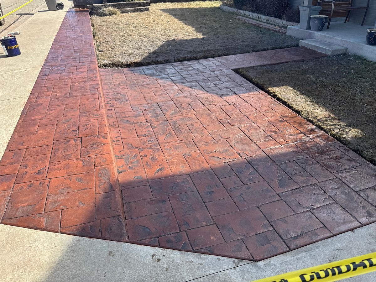 Red brick walkway bordered by grass and concrete, leading past a building entrance.