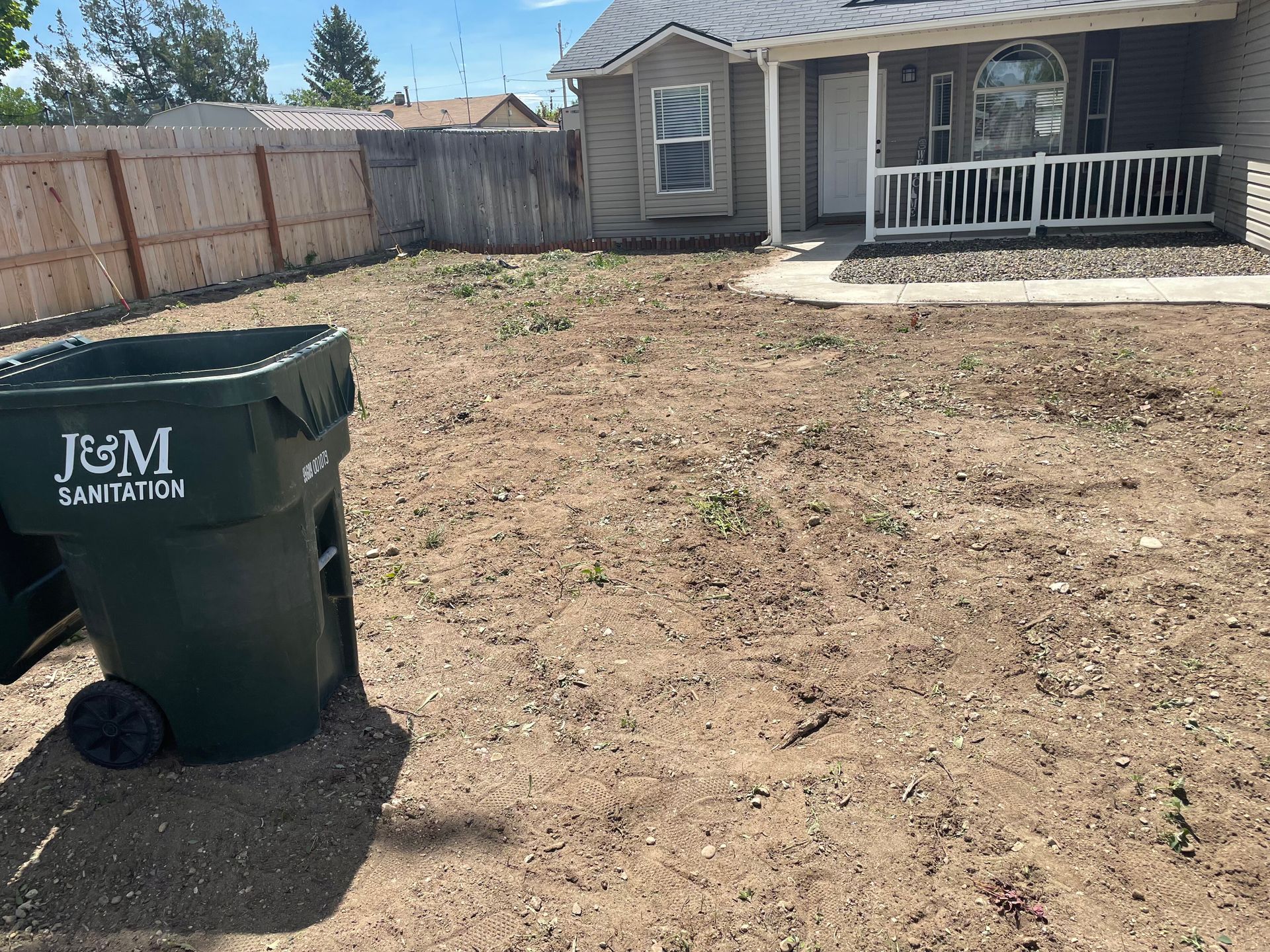 Backyard with dry dirt lawn, trash bin, fenced yard, and a house porch in the background