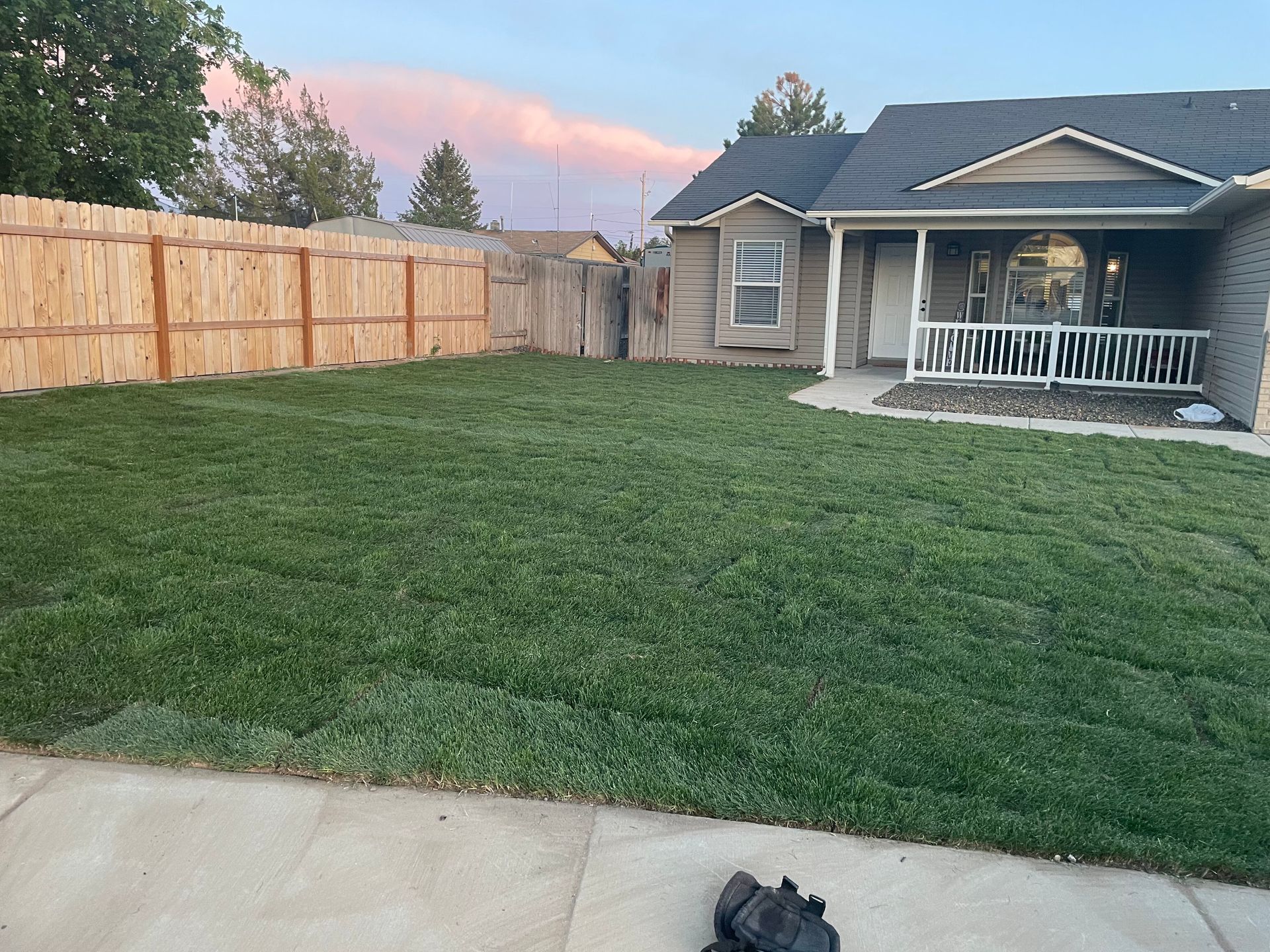 Backyard with green lawn, wooden fence, and a house porch at dusk