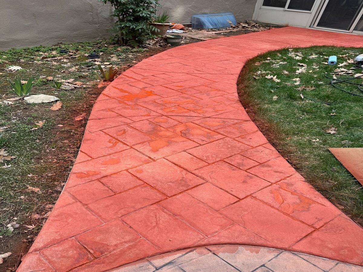 Red brick walkway curving through a yard beside a house and patchy grass.