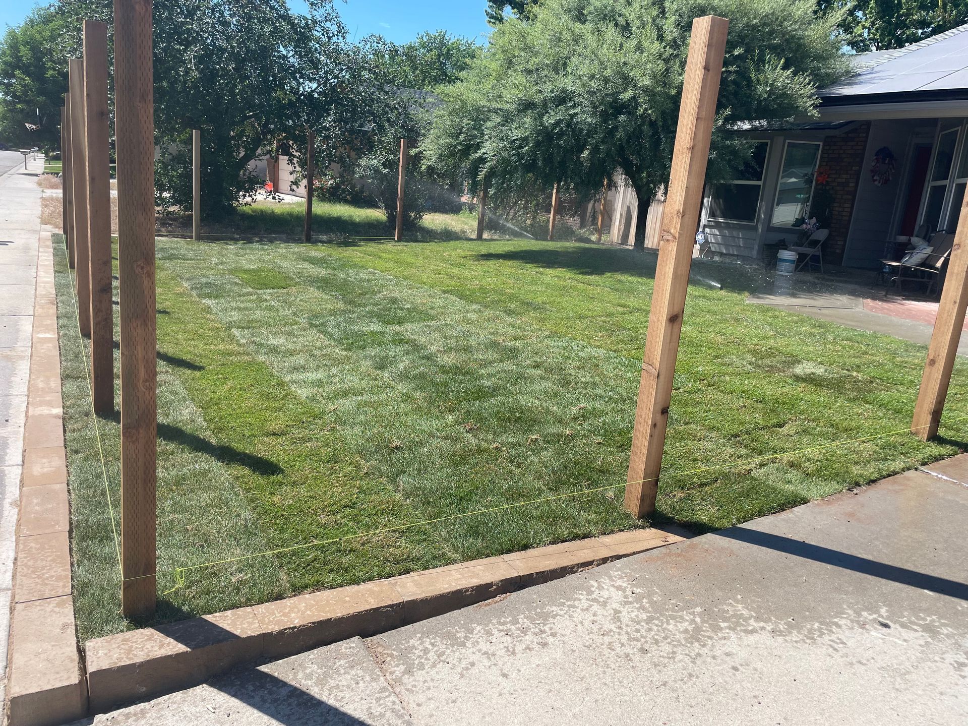 Wooden posts mark an open grassy yard beside a concrete walkway and a building porch.
