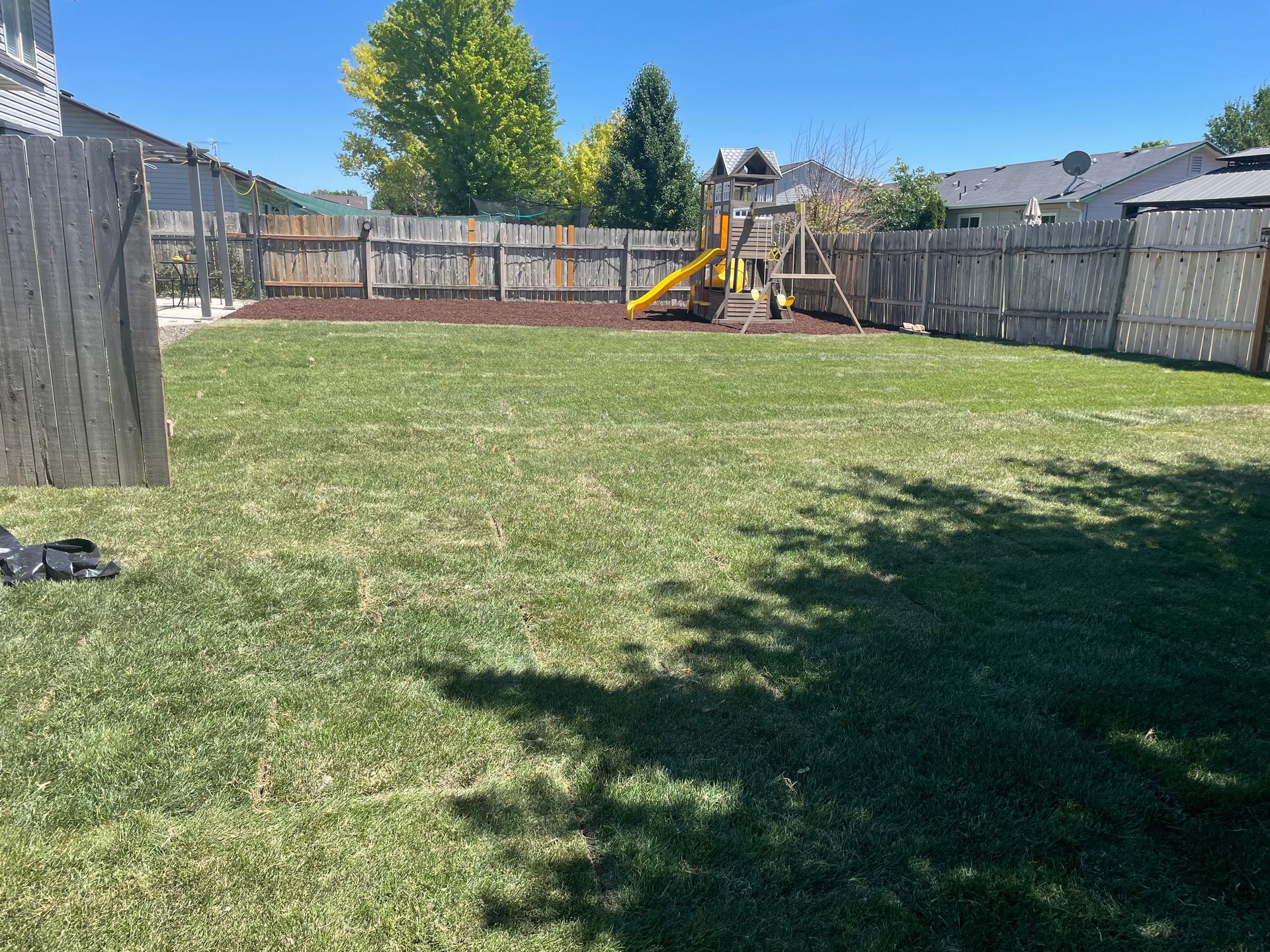 Backyard with a large grassy lawn, wooden fences, and a yellow playground set in the distance.