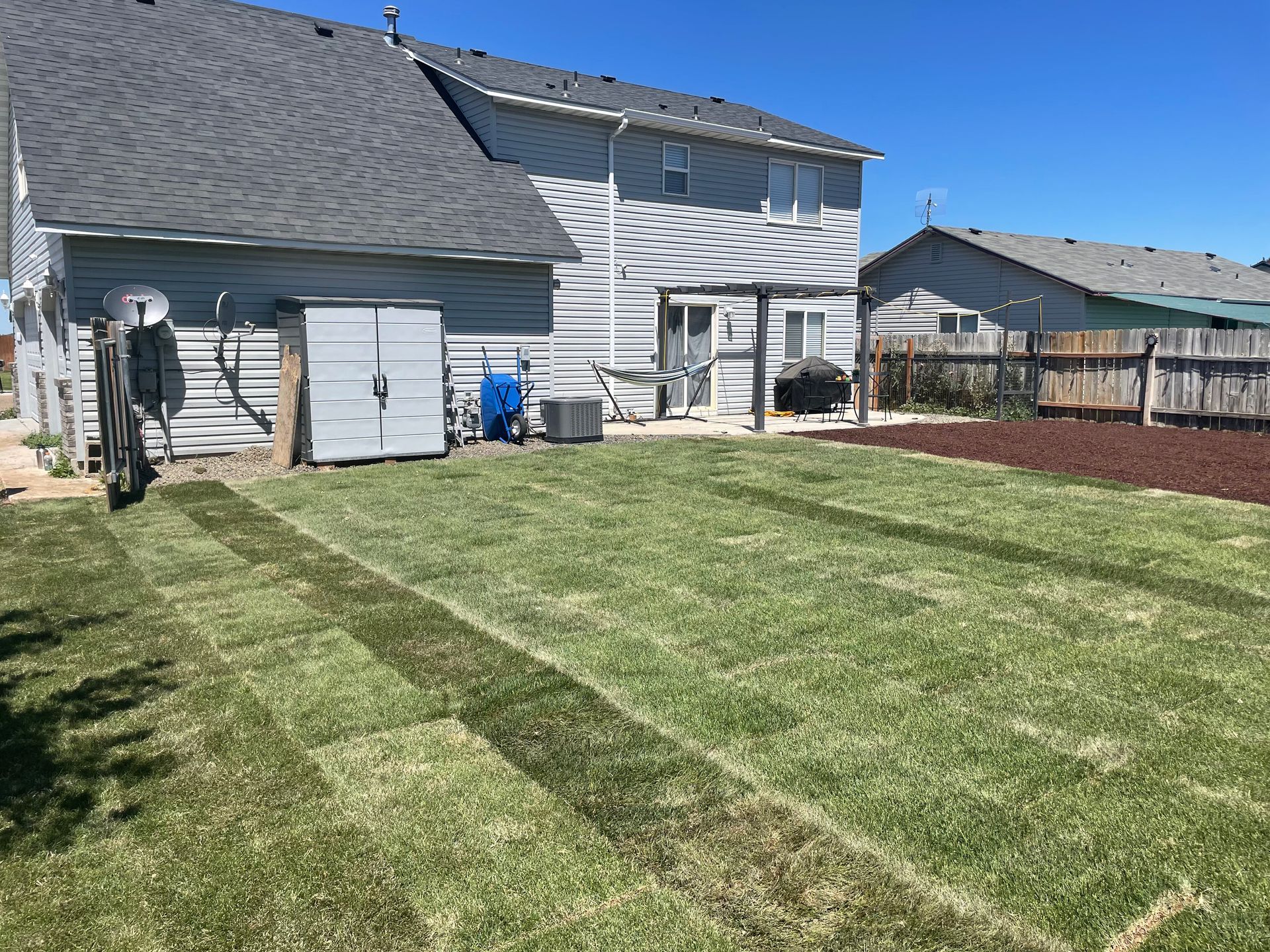 Suburban backyard with freshly mowed lawn, white house, and patio furniture under a blue sky