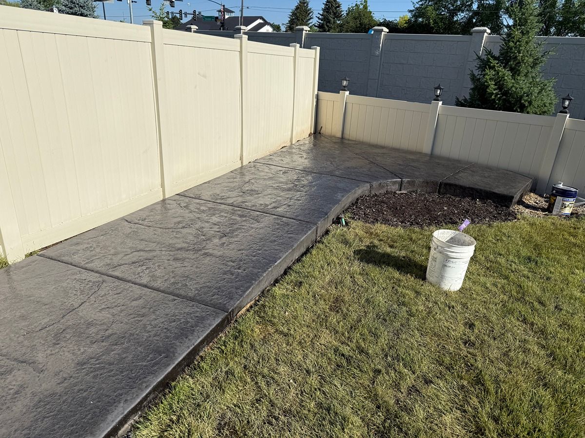 Concrete walkway beside a cream fence, leading to a gate with a grassy yard and white bucket.