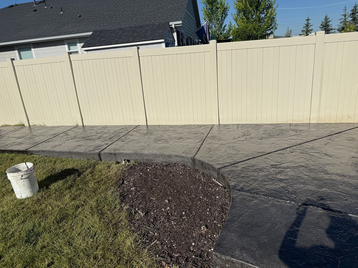 Curved concrete walkway beside a cream fence and grass in a suburban yard, with a white bucket on the lawn
