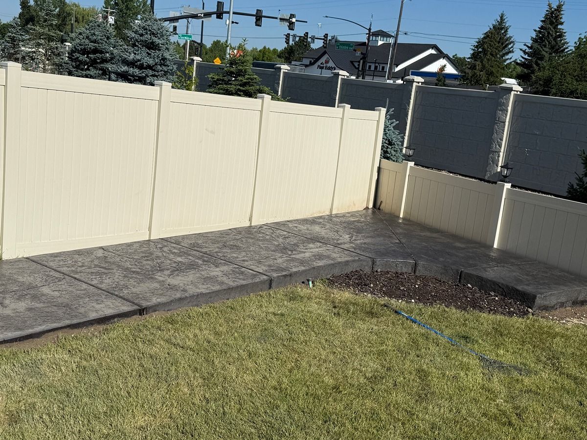 Backyard corner with beige fence, stone patio, and grassy lawn under a clear sky