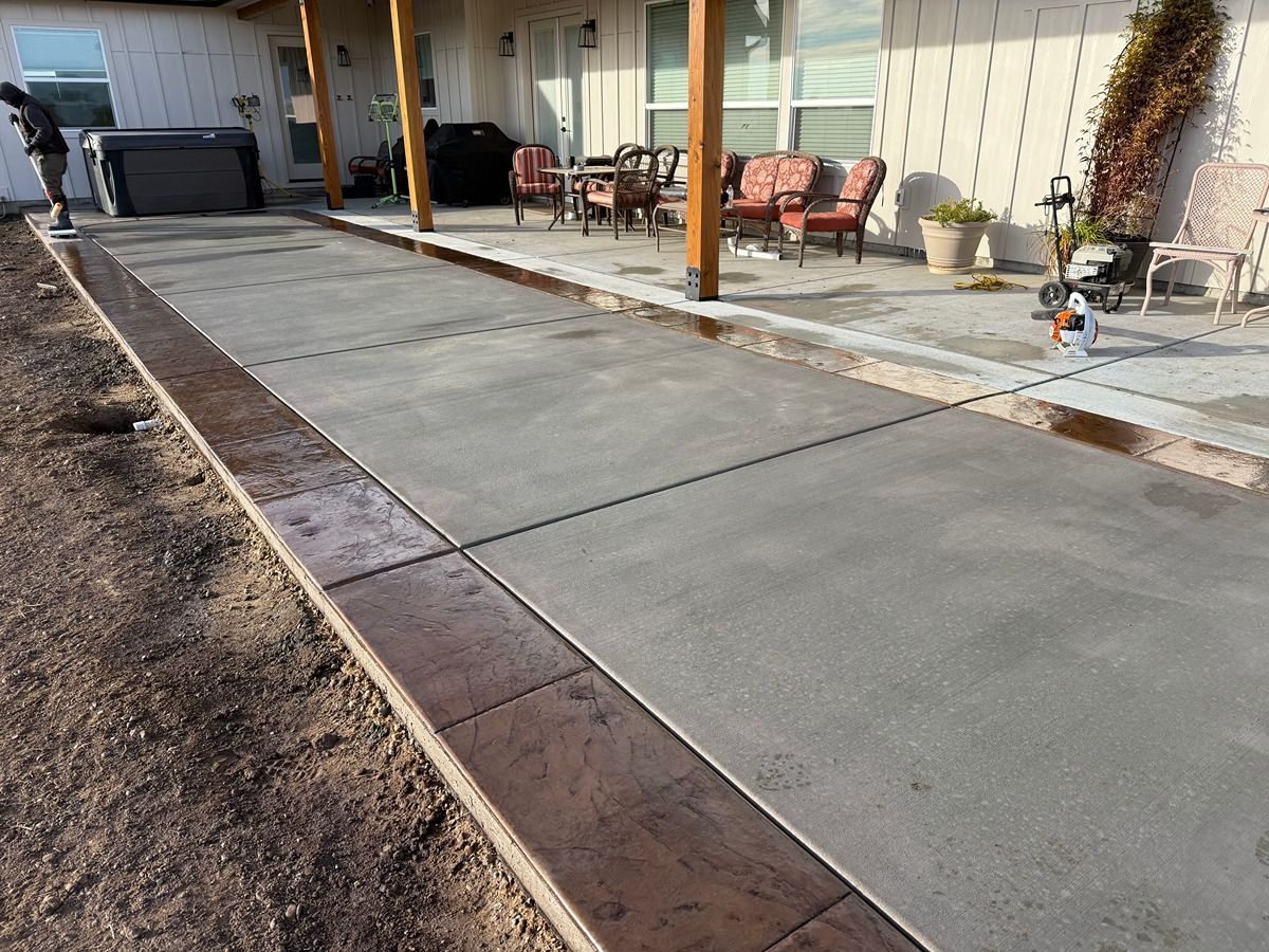 Concrete walkway beside a modern building with outdoor seating and a dirt strip along the left edge
