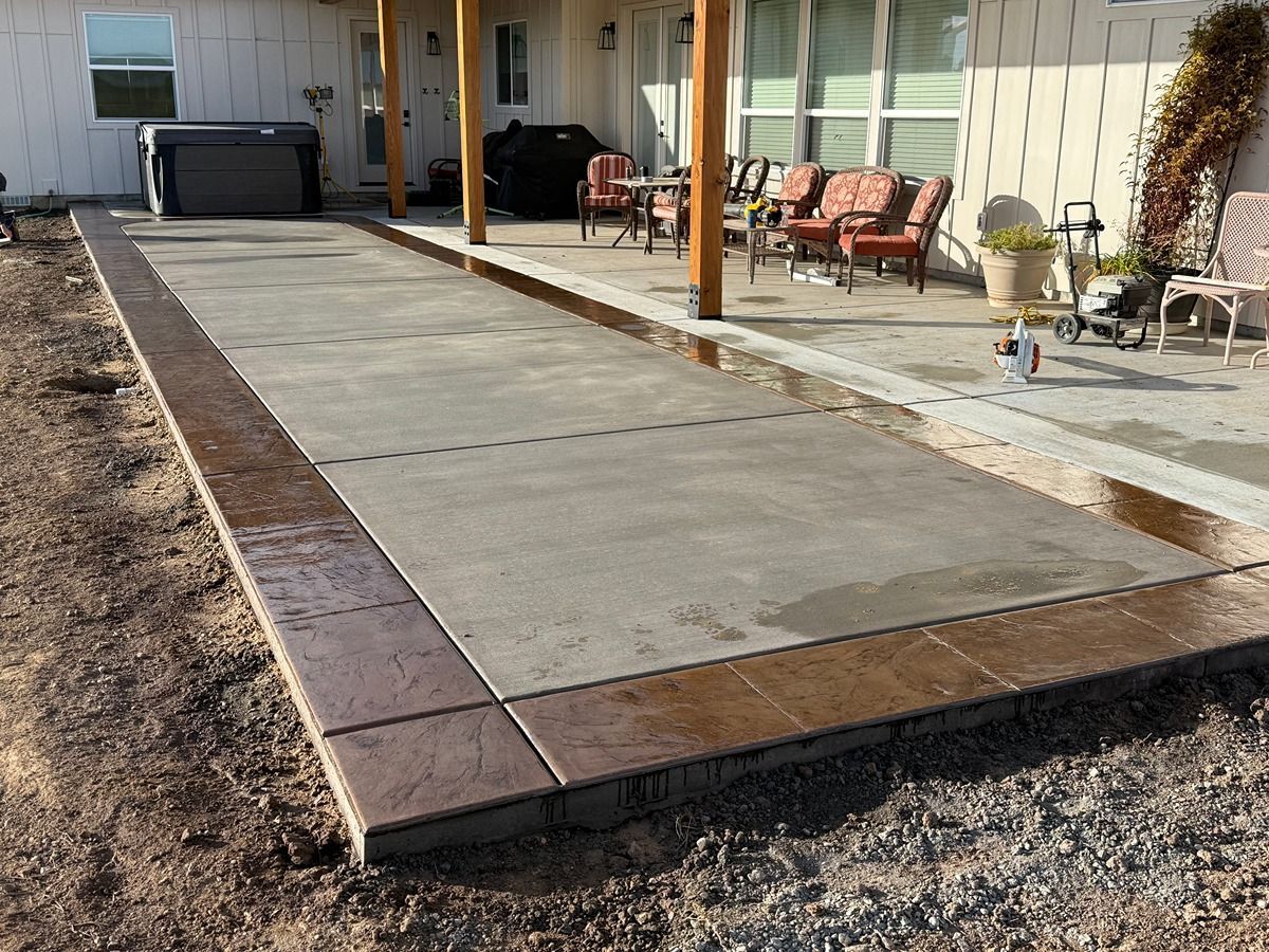 Concrete patio walkway beside a house with brown posts, outdoor chairs, and a grill in the background