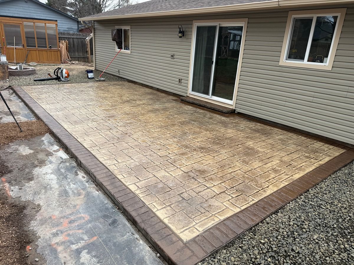 Backyard patio with tan pavers, brick border, and sliding glass door beside a house