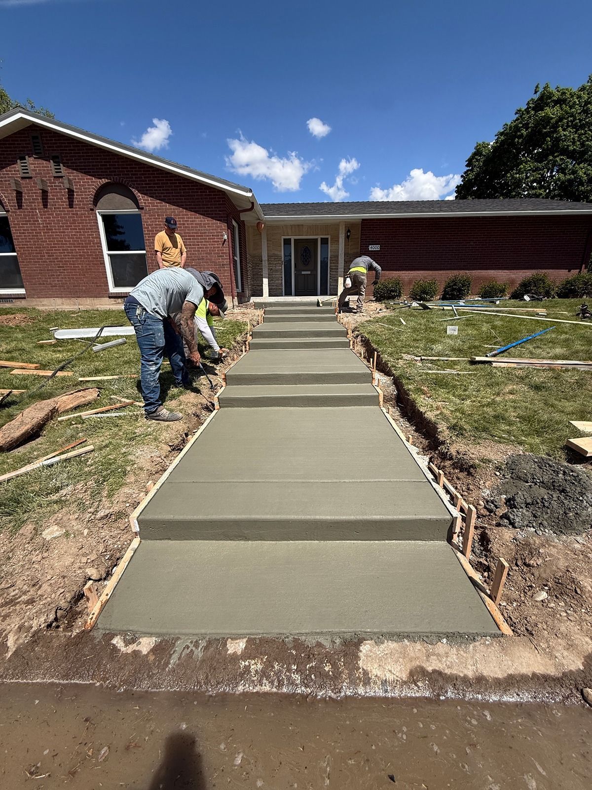 Workers pouring a concrete walkway beside a brick building under a blue sky.