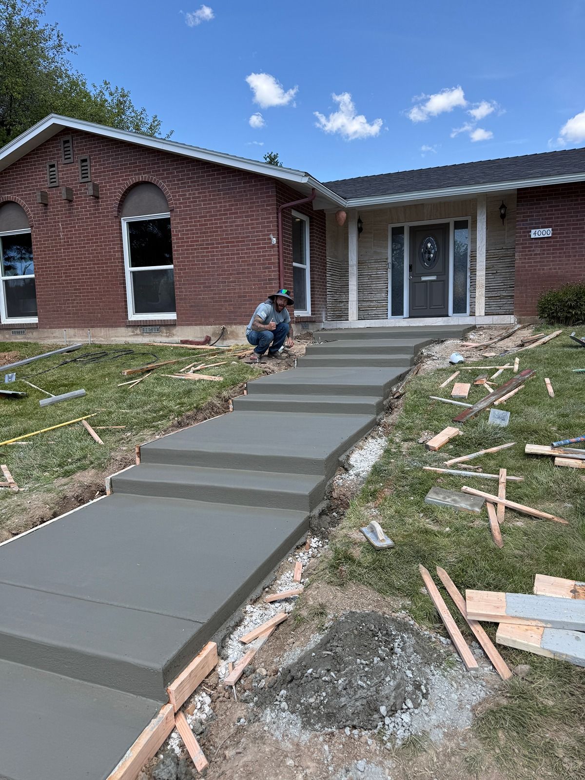 Front walkway under construction leading to a brick house, with a worker smoothing fresh concrete steps.
