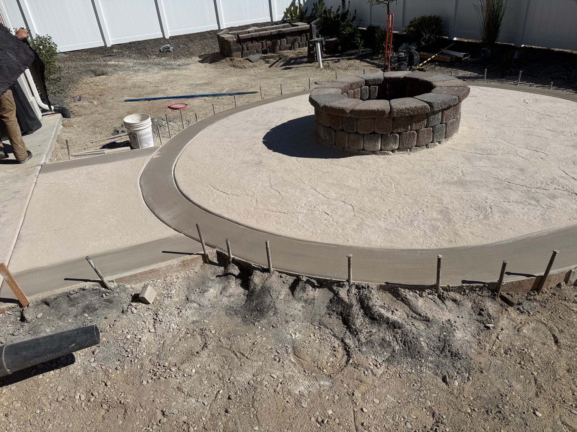 Circular stone fire pit in a gravel courtyard under construction, with concrete walkway and exposed rebar edges