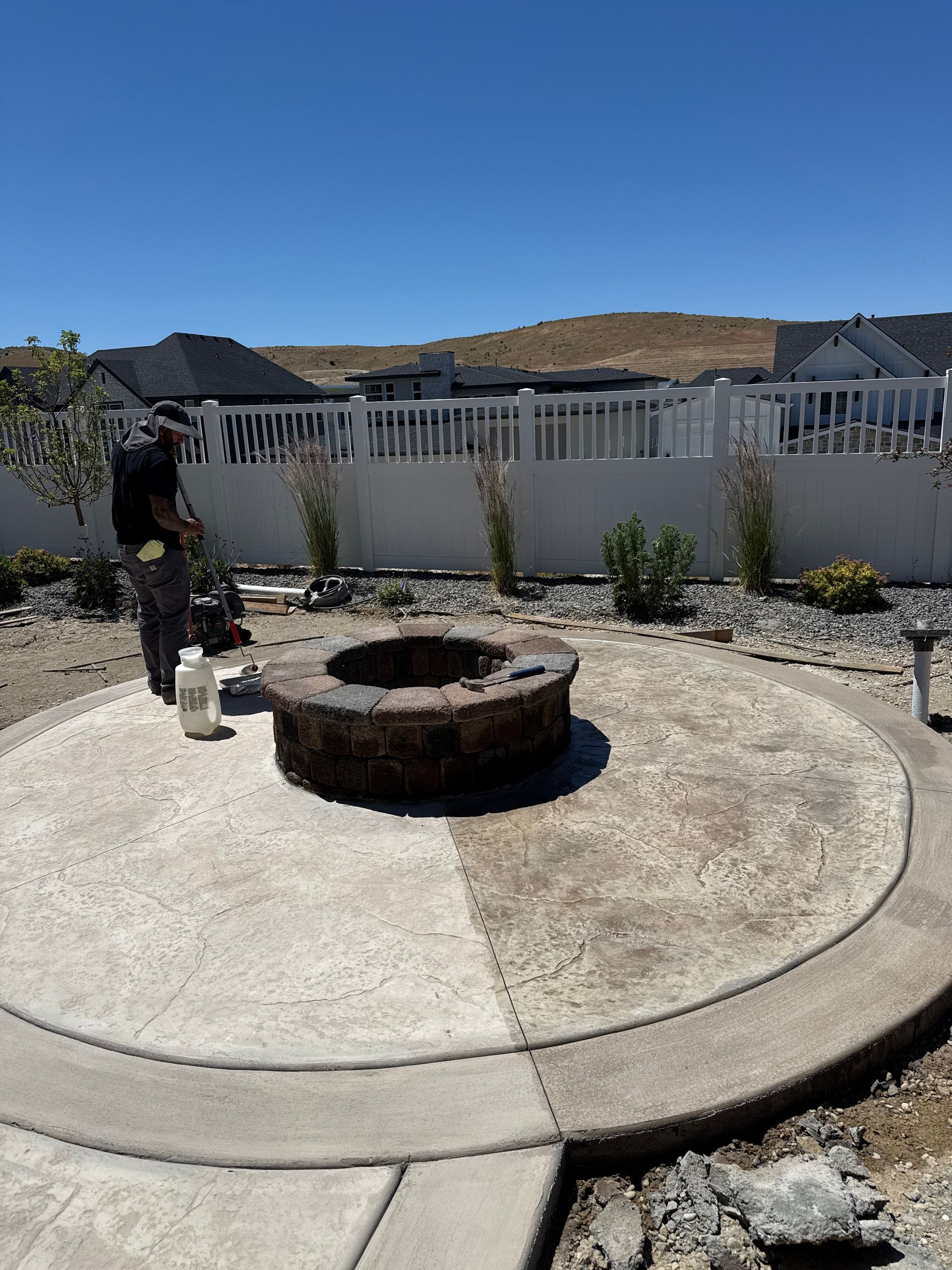 Backyard with circular concrete fire pit and patio beside a white fence under a blue sky