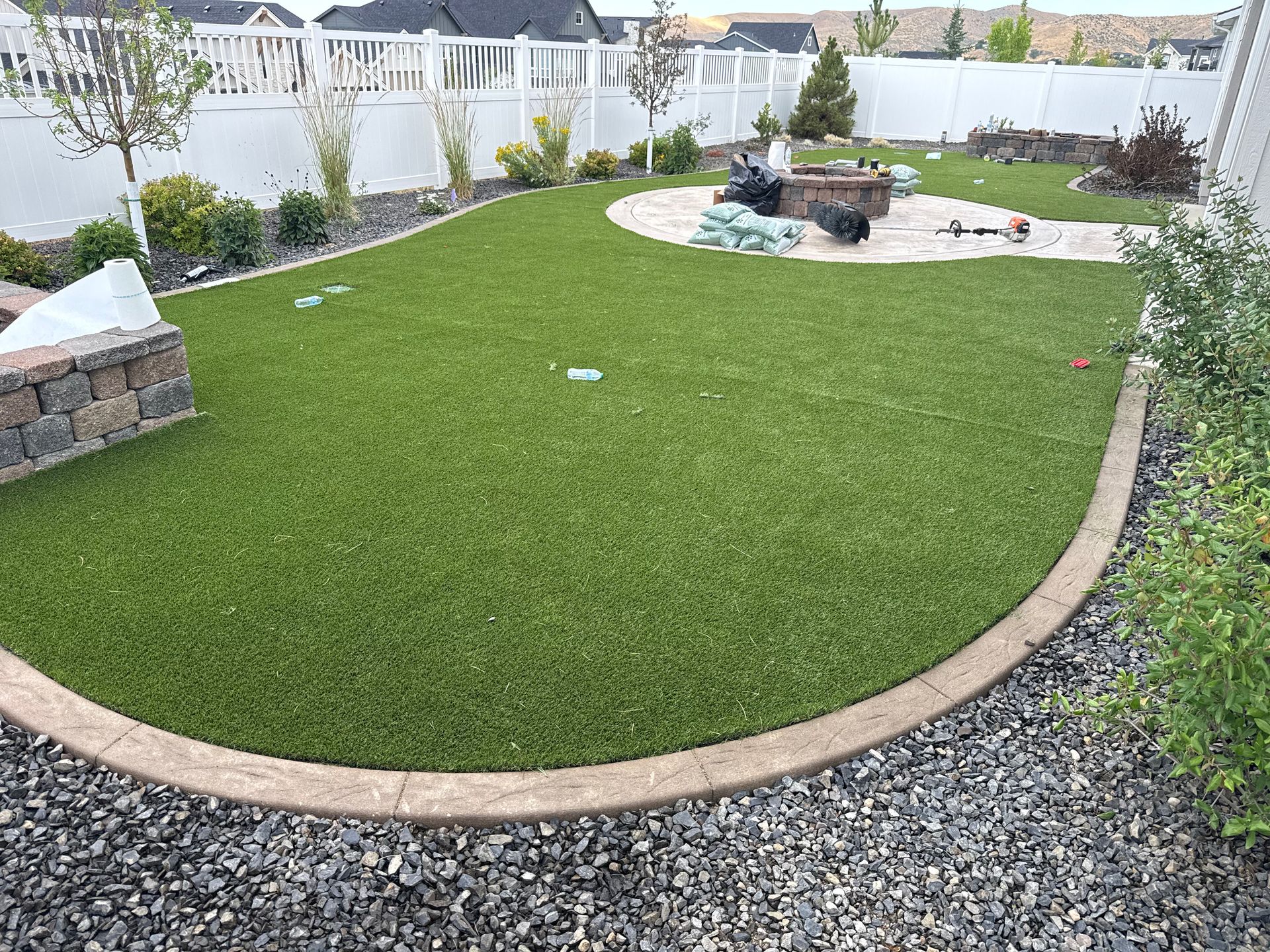 Curved artificial grass lawn with gravel border, stone edging, and a fire pit in a fenced backyard.