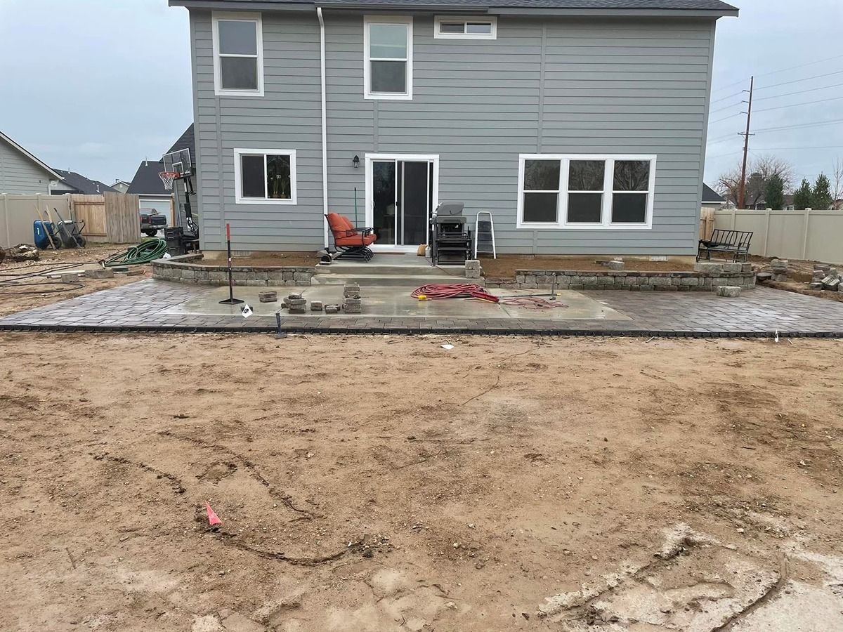 Backyard view of a gray house with a concrete patio, muddy yard, and outdoor furniture near the wall