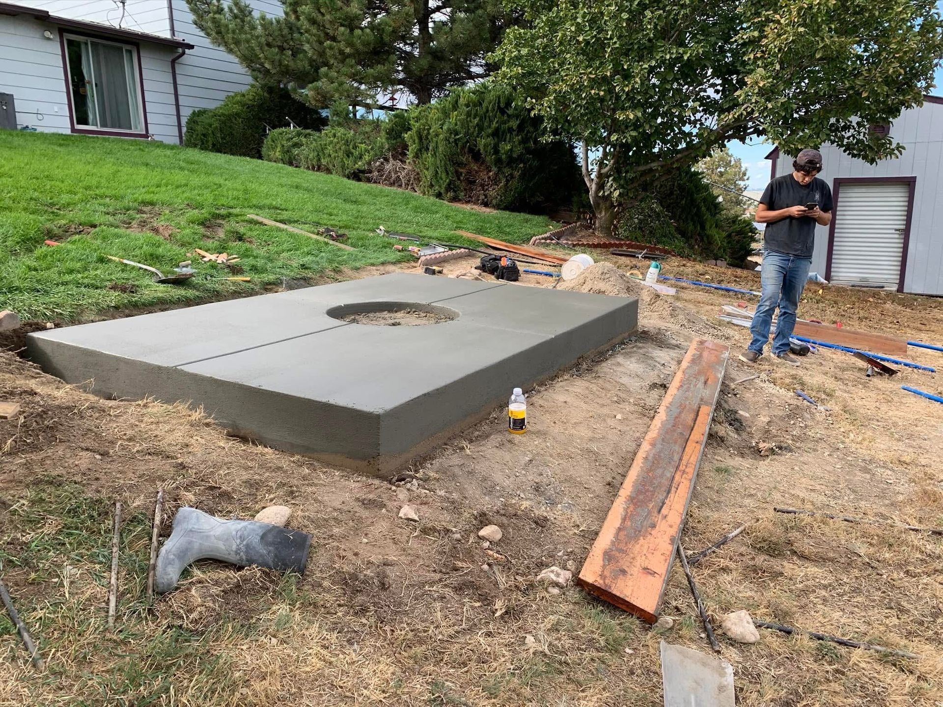 Concrete pad poured in a yard, with a worker standing beside it and lumber nearby.