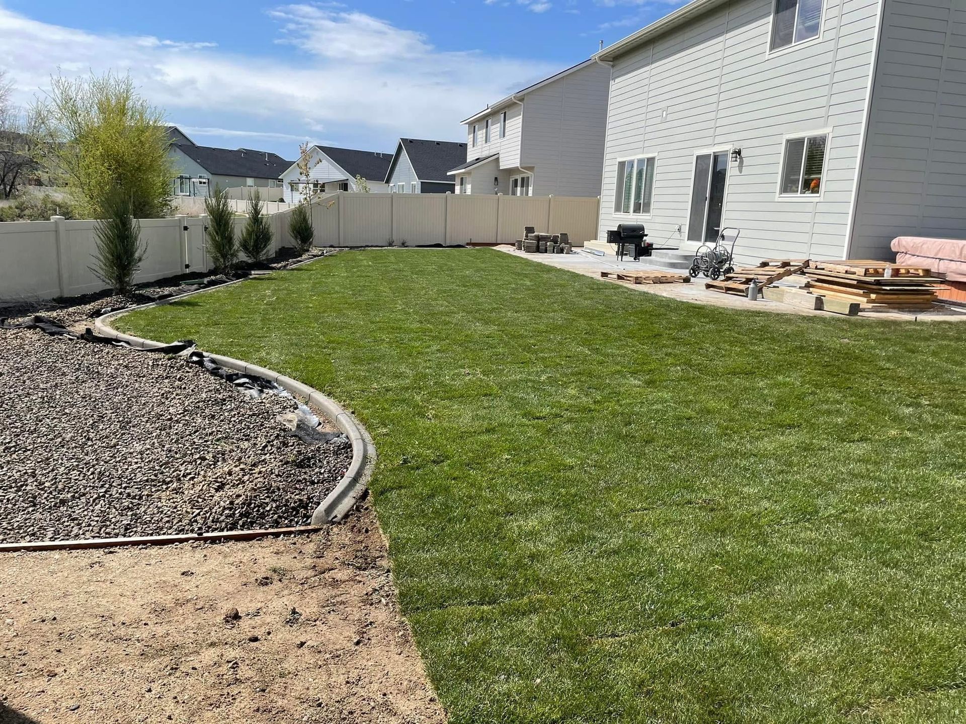 Backyard with green lawn, gravel bed, and patio beside a light-colored house under a blue sky