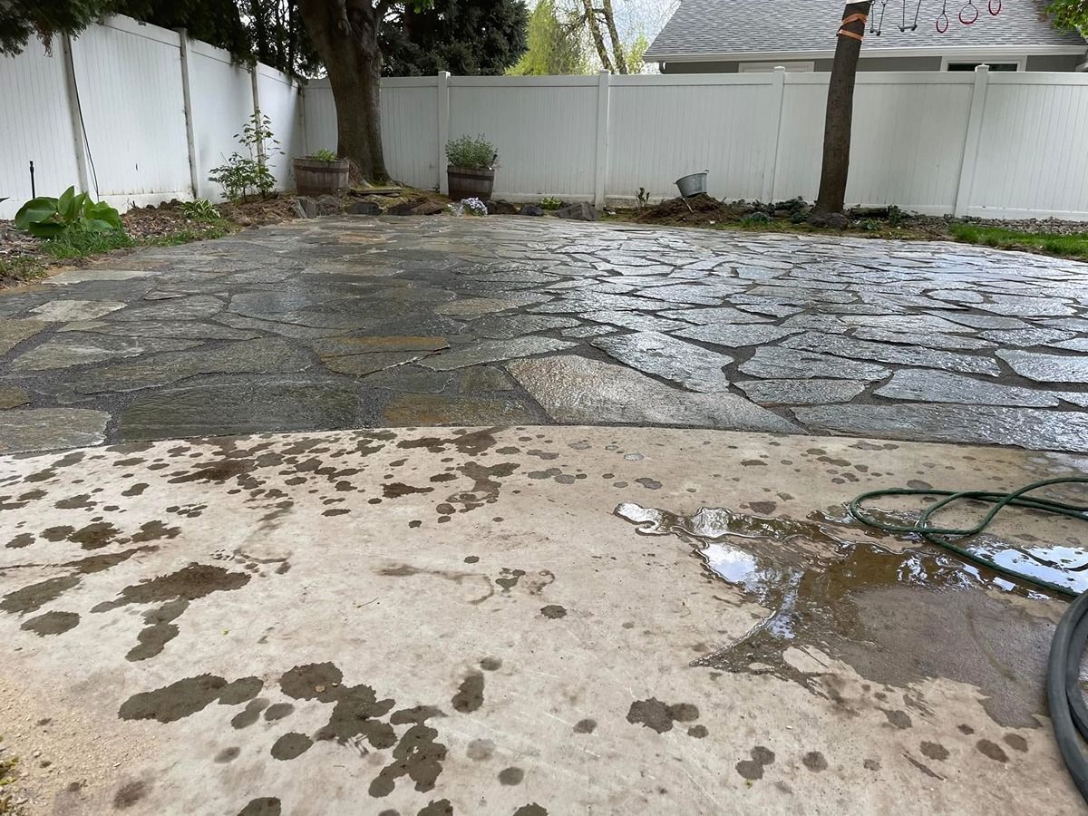 Wet concrete and stone patio in a backyard with puddles, hose, and white fence.