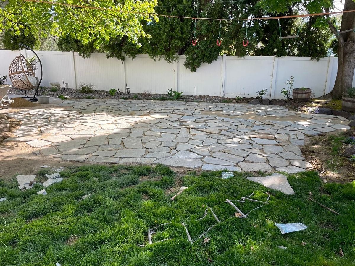 Stone patio in a backyard with green grass, white fence, and trees casting shade