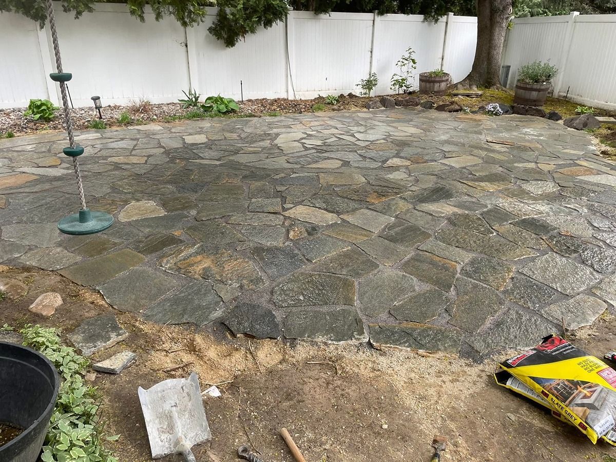 Stone patio under construction in a backyard, with scattered soil, tools, and a white fence along the edge