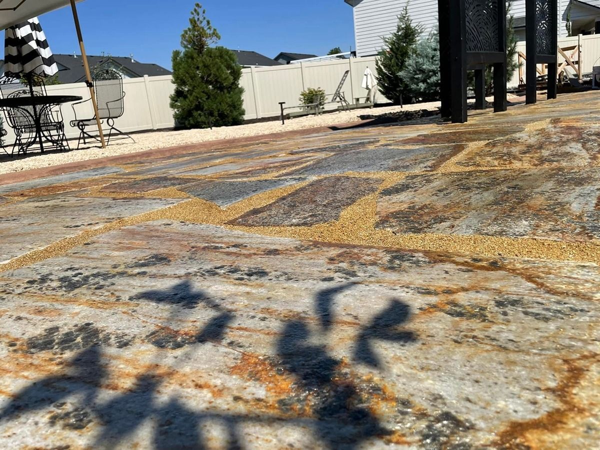 Sunlit patio with patterned concrete, shadows, a metal table, and chairs near a building and trees