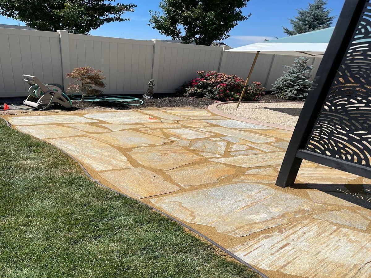 Backyard stone patio with lawn, fence, potted plants, and a black metal chair edge