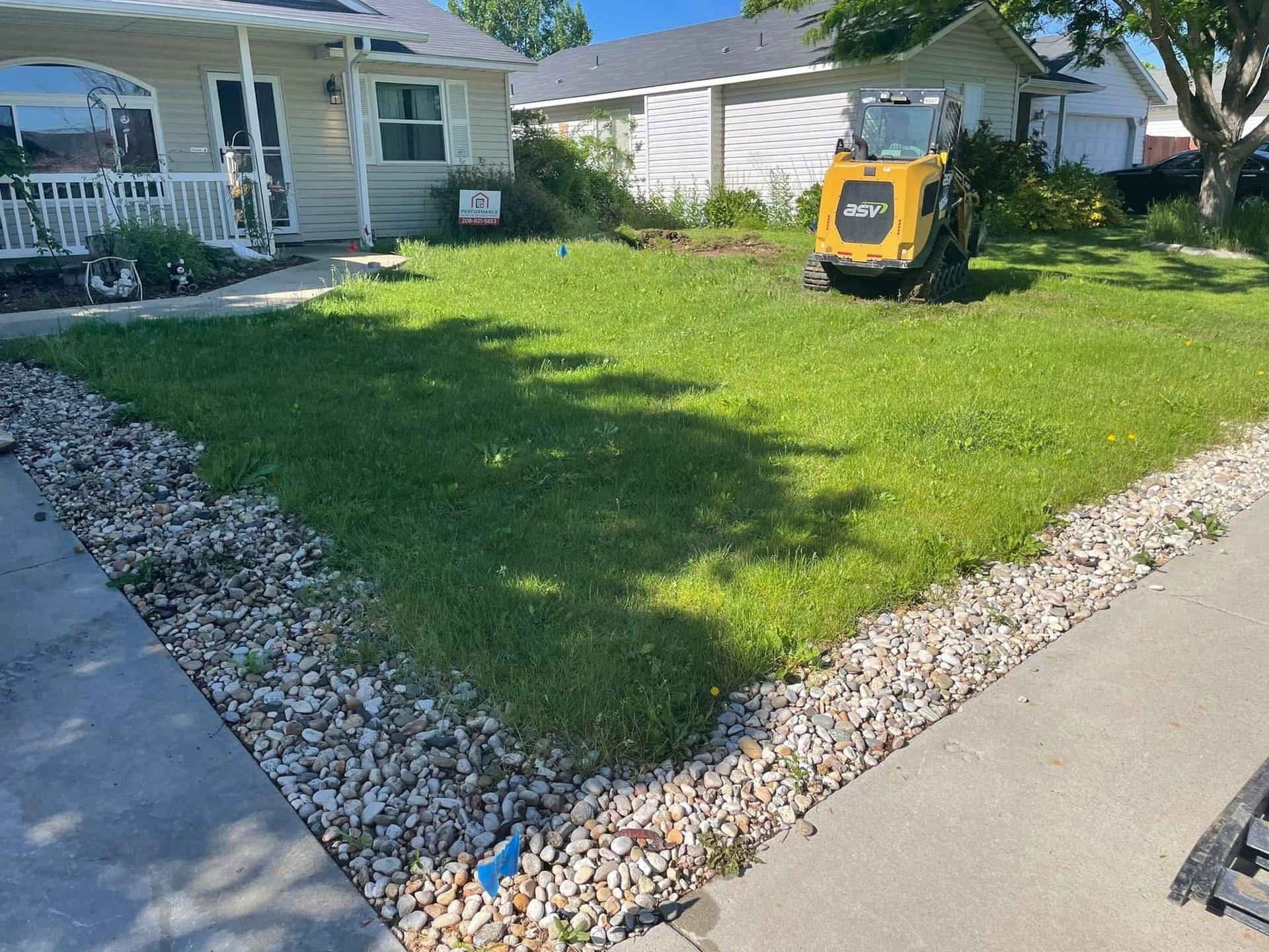 Small grassy front yard bordered by rocks, with a yellow lawn mower and houses in the background