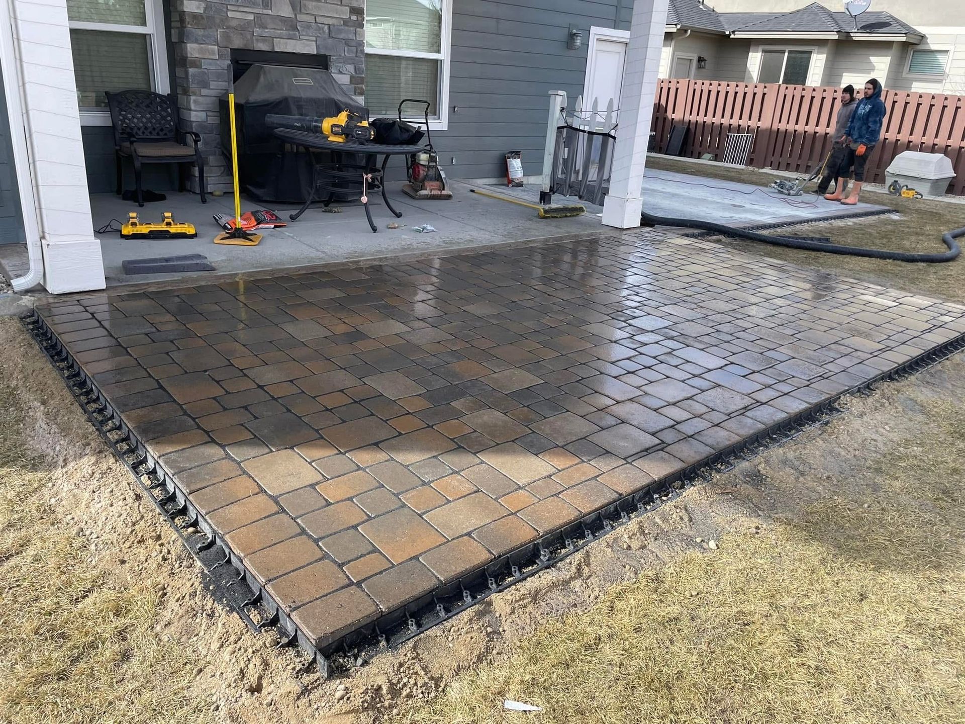 Freshly laid brick patio beside a house, with workers finishing the edges and a lawn nearby