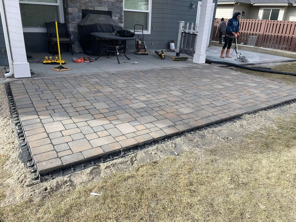 Back patio with gray paver deck, grill, and two people working near the house