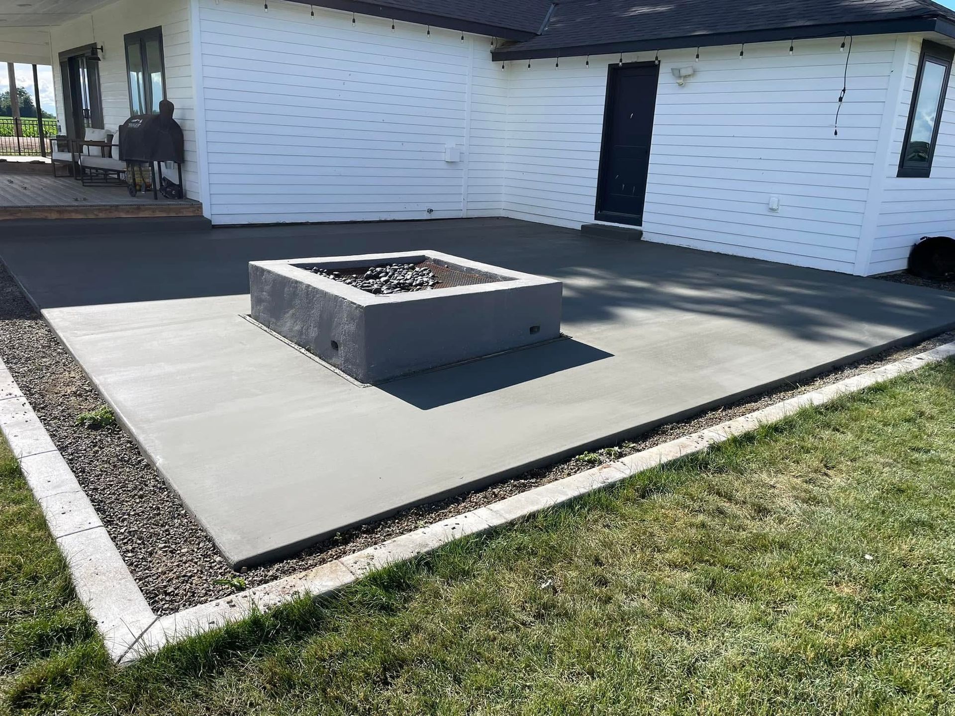 Concrete patio with a square fire pit beside a white house and grassy yard.
