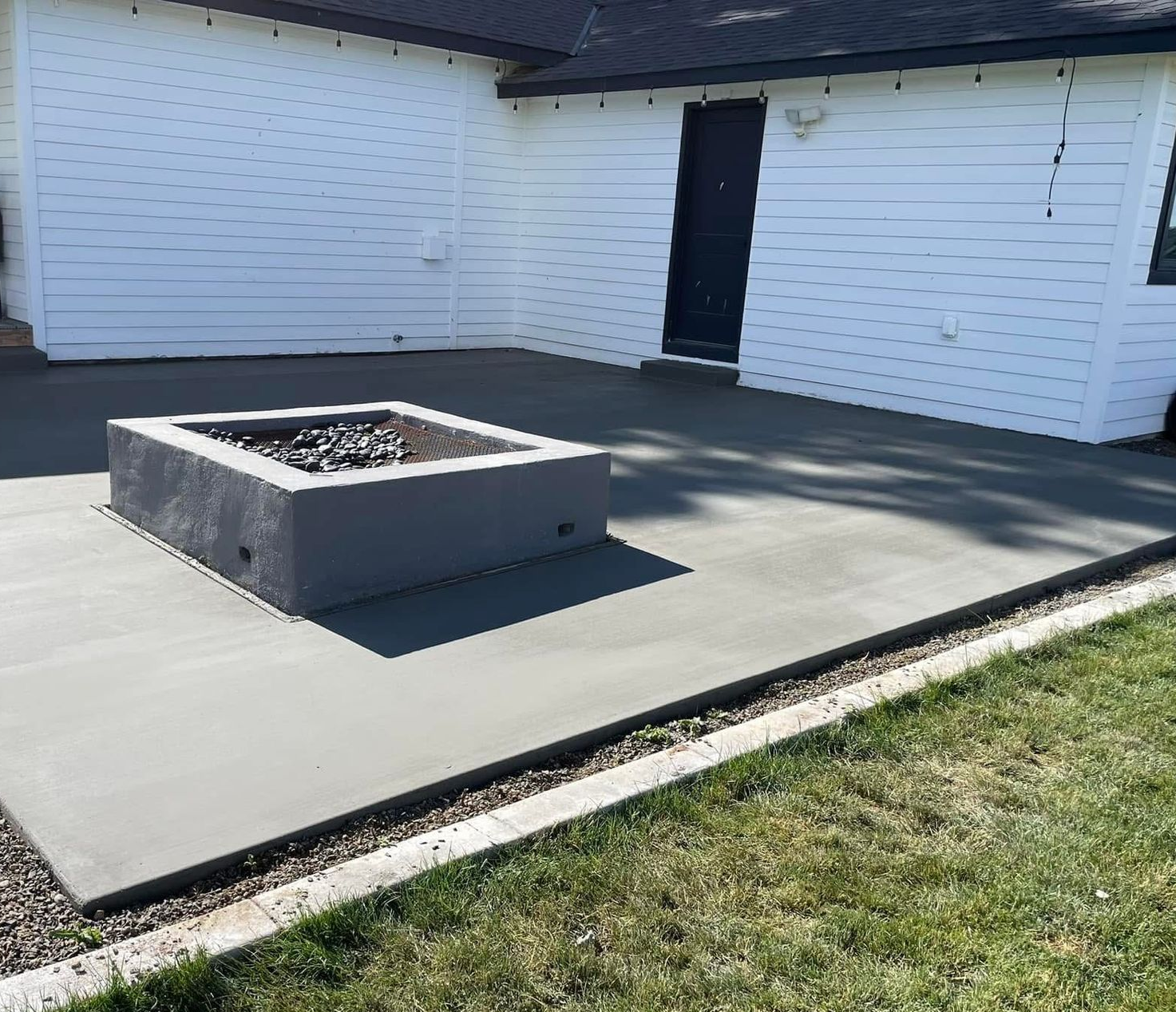 Concrete patio with a square fire pit beside a white building and a patch of grass
