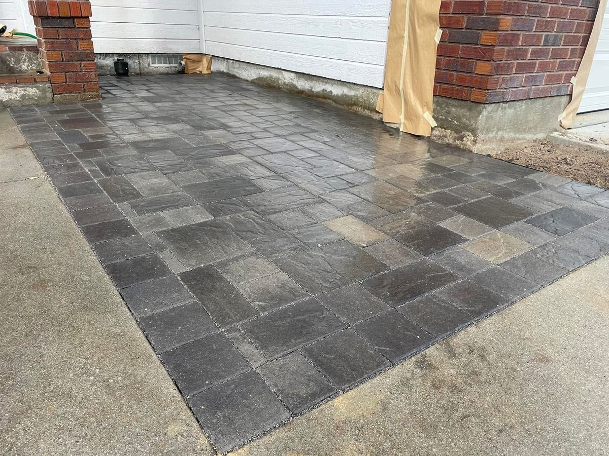 Newly laid dark gray paver patio beside a house with brick pillars and white siding