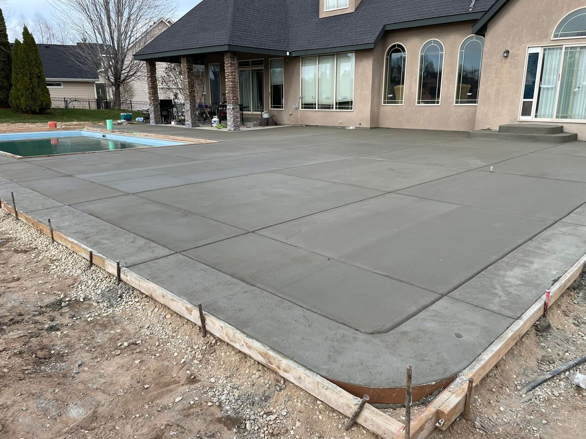 Freshly poured concrete patio beside a house under construction, with foundation and pool area visible