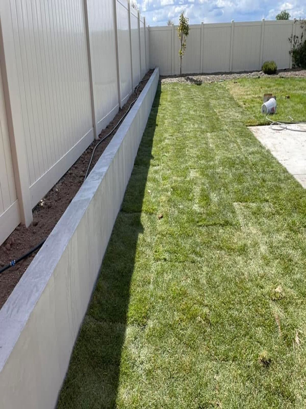 Backyard with green lawn, concrete retaining wall, and white fence under a partly cloudy sky